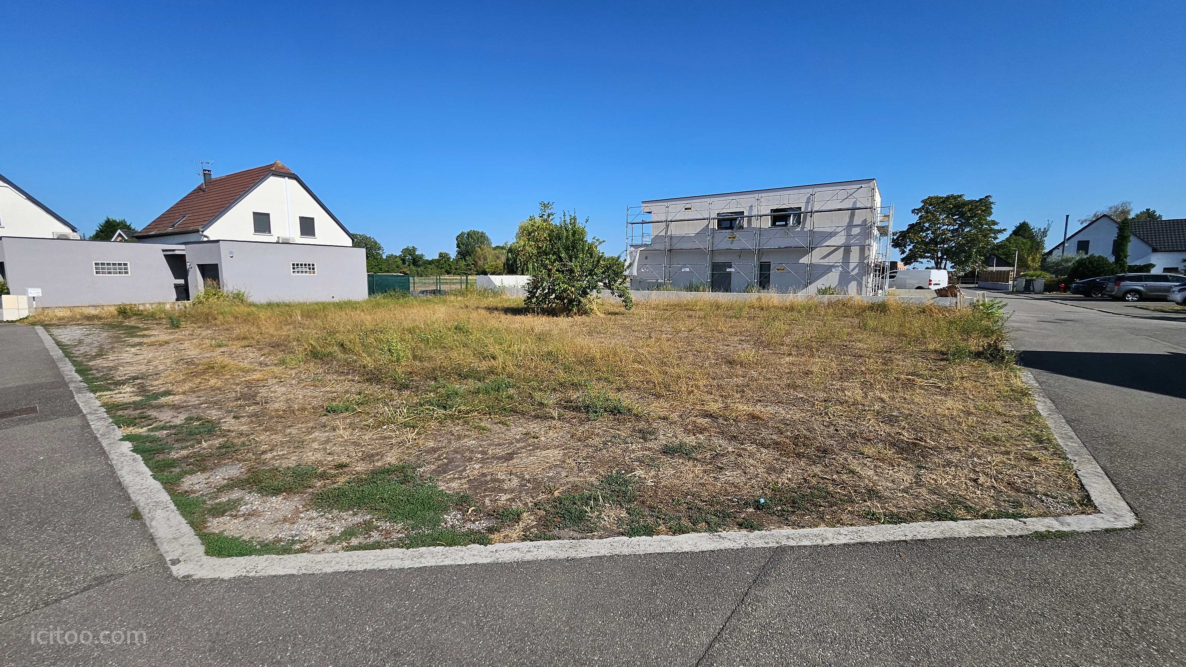 Terrain constructible à vendre à Jebsheim, avec des maisons voisines et un ciel bleu.